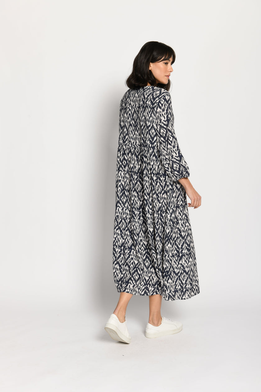 Model trägt luftiges Stufenkleid mit langen Ärmeln und tollem Print in den Farben Weiß und Dunkelblau – Rückansicht