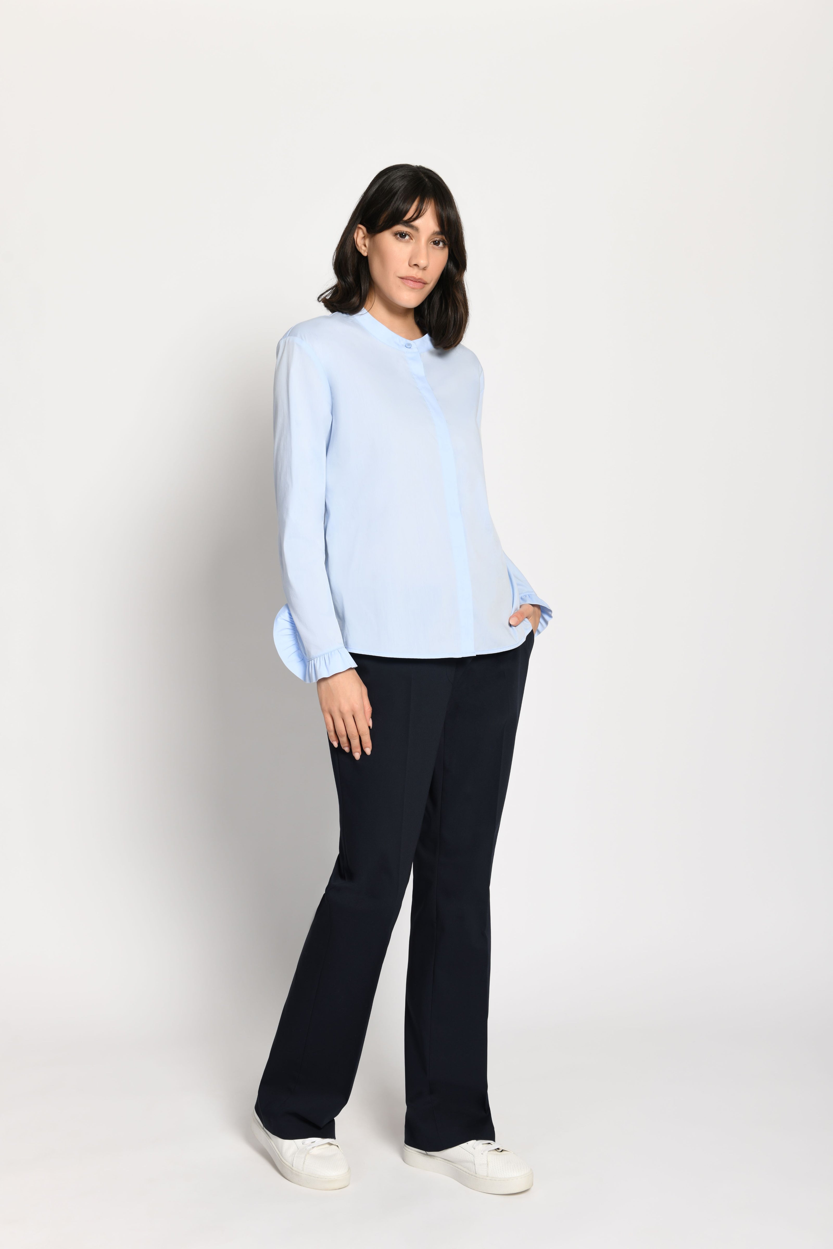 Model trägt hellblaue, locker sitzende Damenbluse mit Rüschen an den Ärmeln – seitliche Pose, Blick nach vorne