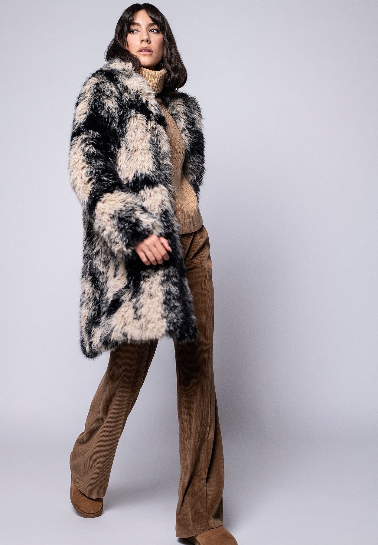 Damenmantel aus langhaarigem schwarz-beigen Webpelz lässig kombiniert zu brauner Cordhose und Rollkragenpullover.